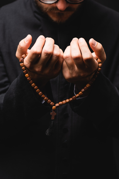 cropped view of pastor holding rosary beads in hands and praying isolated on black  - Zdjęcie, obraz
