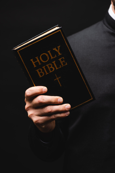 cropped view of priest holding holy bible in hand isolated on black  - Fotografie, Obrázek