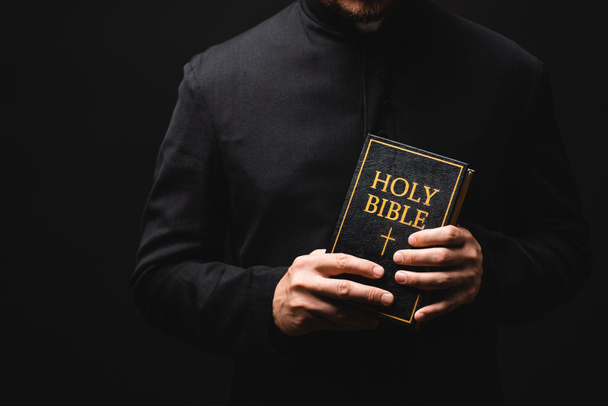 обрезанный вид священника, держащего в руках святую Библию, изолированную на черном  - Фото, изображение