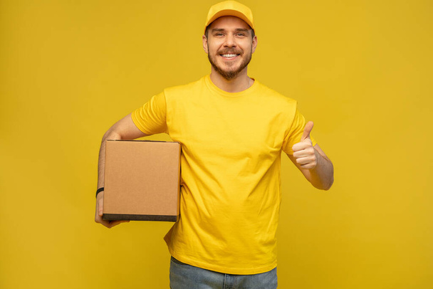 Portrait of excited delivery man in yellow uniform holding paper box isolated over yellow background. - Фото, зображення