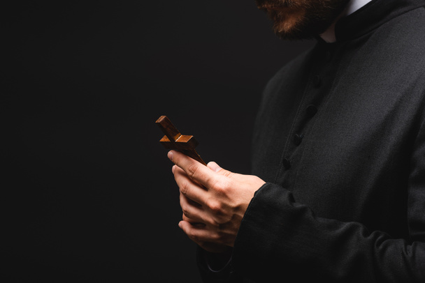 καλλιεργημένη άποψη του ιερέα που κρατά το σταυρό απομονωμένο σε μαύρο  - Φωτογραφία, εικόνα