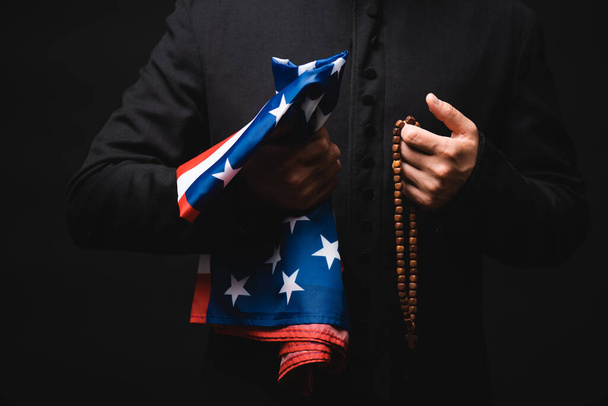 黒で隔離されたアメリカ国旗とバラ色のビーズを手に持つ司祭のクロップドビュー  - 写真・画像