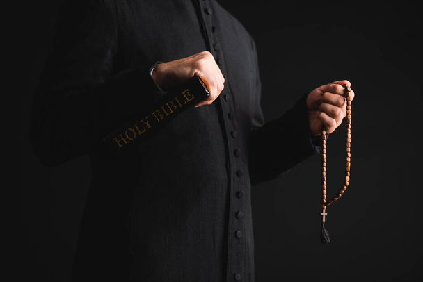 vista ritagliata di pastore che tiene santa Bibbia e rosario perline in mani isolate su nero  - Foto, immagini