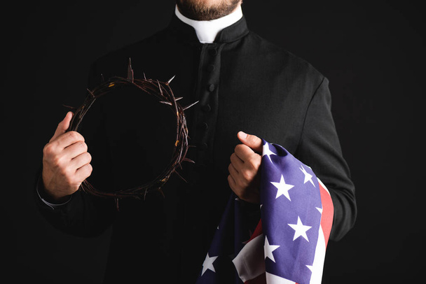 vista ritagliata del sacerdote che tiene corona con punte e bandiera americana isolata su nero  - Foto, immagini