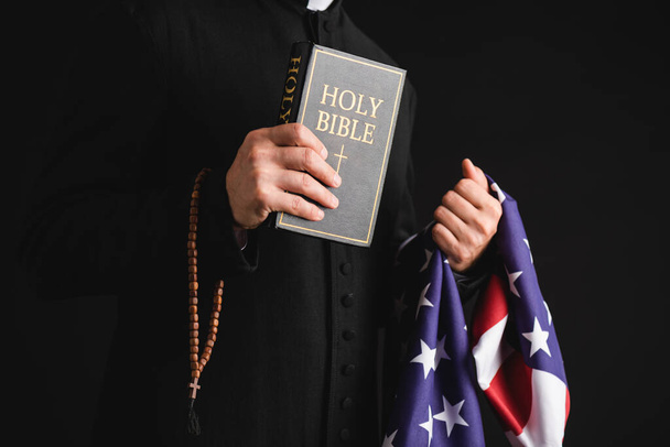 vista cortada de padre segurando bíblia sagrada, bandeira americana e rosário contas isoladas em preto
  - Foto, Imagem