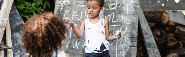 panoramic shot of poor african american kid standing near chalkboard and curly sister  - Photo, Image