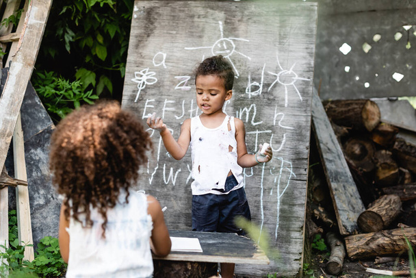 enfoque selectivo de niño afroamericano pobre de pie cerca de pizarra con letras y hermana rizada
 - Foto, imagen