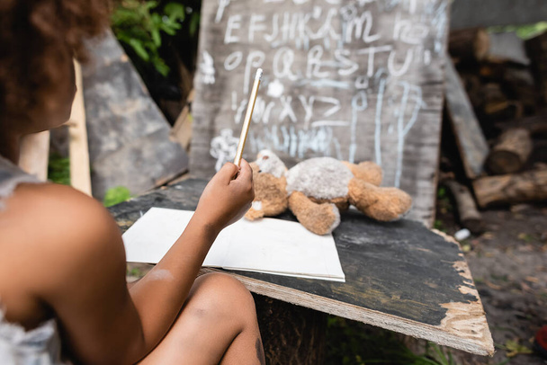 partial view of african american child holding pencil near teddy bear and chalkboard  - Фото, зображення