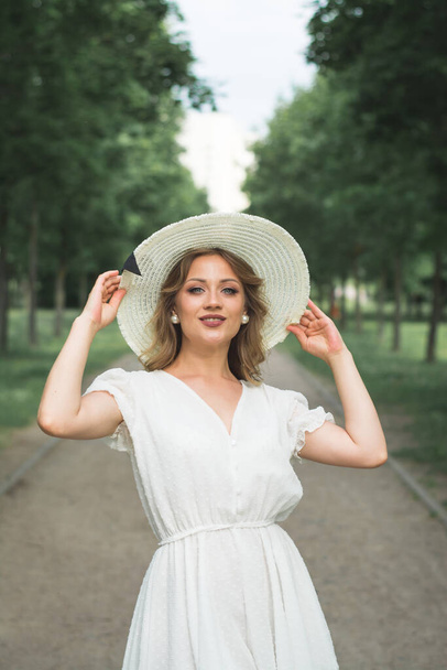 Menina bonita elegante em um chapéu de verão branco e um grande chapéu branco posa para um fotógrafo em um caminho em um parque de verão
 - Foto, Imagem