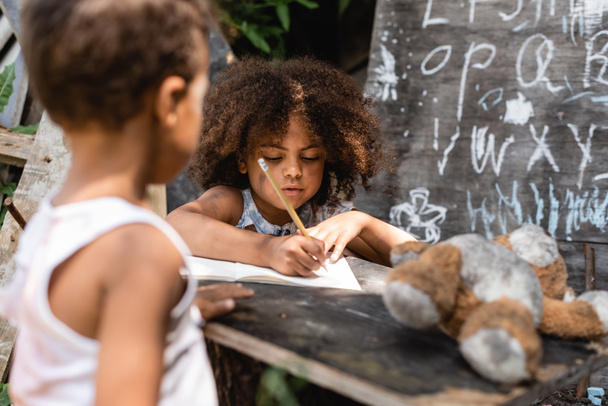 Selektiver Fokus armer afrikanisch-amerikanischer Kinder, die in der Nähe ihres Bruders auf Papier schreiben - Foto, Bild