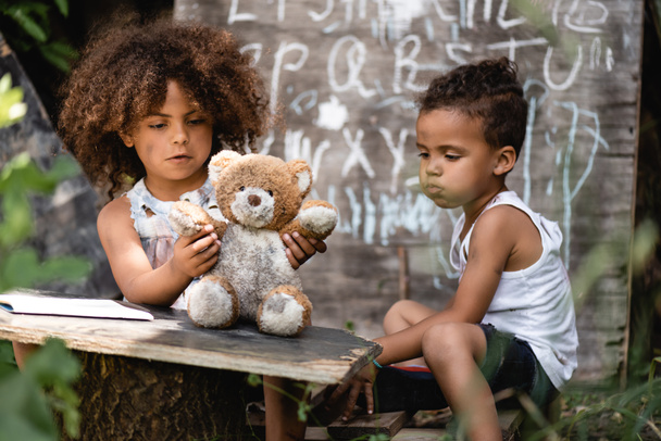 selective focus of poor african american kid playing with dirty teddy bear near brother - Foto, afbeelding