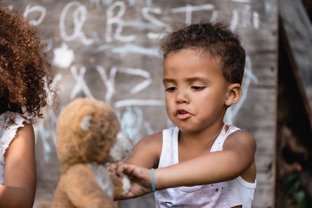 selective focus of poor african american boy playing with dirty teddy bear near child - Fotografie, Obrázek