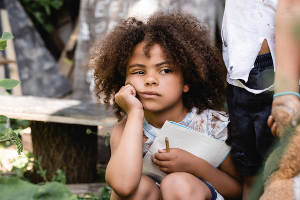 φτωχό αφροαμερικανό παιδί κρατώντας σημειωματάριο κοντά στο παιδί στέκεται με βρώμικο μαλακό παιχνίδι - Φωτογραφία, εικόνα