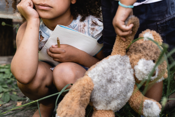 abgeschnittene Ansicht eines armen afrikanisch-amerikanischen Kindes, das ein Notizbuch in der Nähe eines Kindes hält, das mit schmutzigem Stofftier steht - Foto, Bild