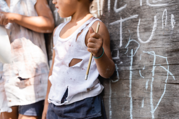 обрезанный вид бедного африканского американского ребенка в порванной одежде, стоящего рядом с доской и сестрой, держа карандаш  - Фото, изображение