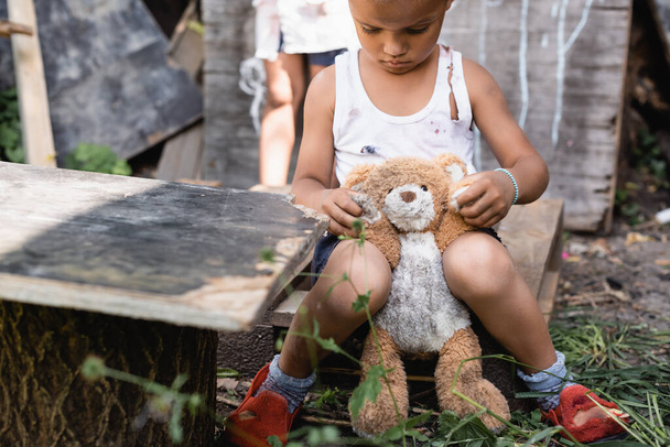 selectieve focus van arme Afrikaanse Amerikaanse kind in gescheurde kleren zitten met zacht speelgoed  - Foto, afbeelding