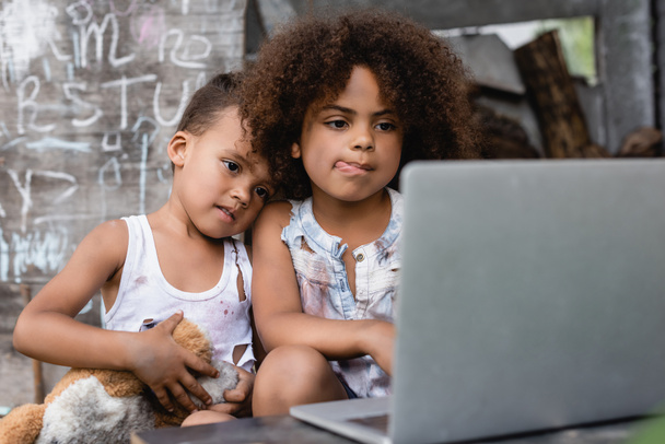 selective focus of poor african american kid sticking out tongue near brother and using laptop outside  - Foto, afbeelding