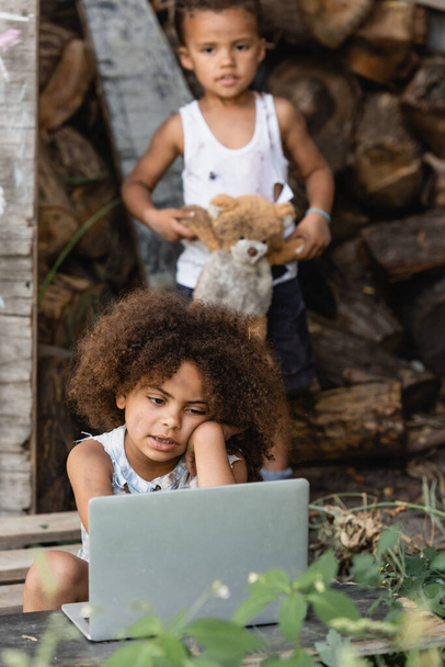 Enfoque selectivo del niño afroamericano sentado cerca de la computadora portátil y hermano con oso de peluche en el barrio pobre
  - Foto, imagen