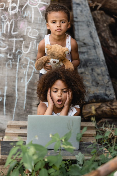 Selective focus of shocked african american child looking at laptop near brother holding teddy bear on urban street  - Photo, Image