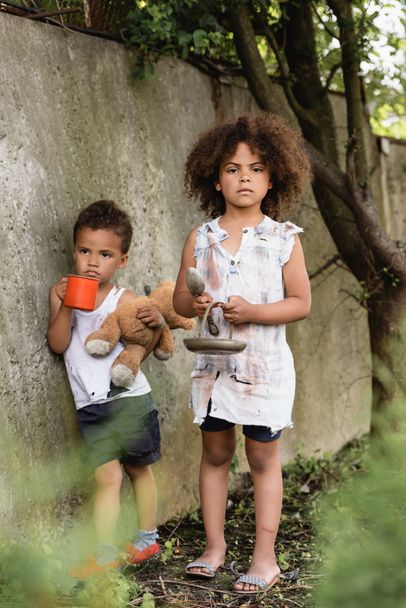 Επιλεκτική εστίαση του φτωχού αφροαμερικανού παιδιού που κρατά μεταλλική πλάκα και κουτάλι κοντά στον αδελφό με κύπελλο και βρώμικο αρκουδάκι κοντά σε τσιμεντένιο τοίχο στην αστική οδό  - Φωτογραφία, εικόνα