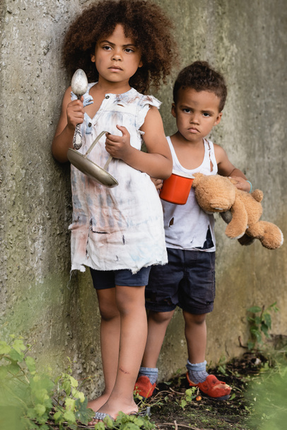 Selectieve focus van dakloze Afrikaans-Amerikaanse kinderen met bord en beker, terwijl bedelen aalmoezen in sloppenwijk  - Foto, afbeelding