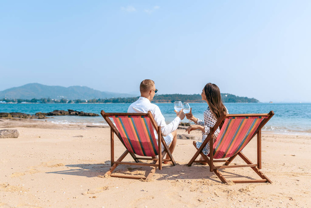 Smiling partners enjoying drinks champagne while resting on comfortable chairs by sea beach. - 写真・画像