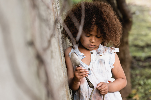 Selectieve focus van arm en boos Afrikaans Amerikaans kind houden vuile metalen lepel en plaat in sloppenwijk  - Foto, afbeelding