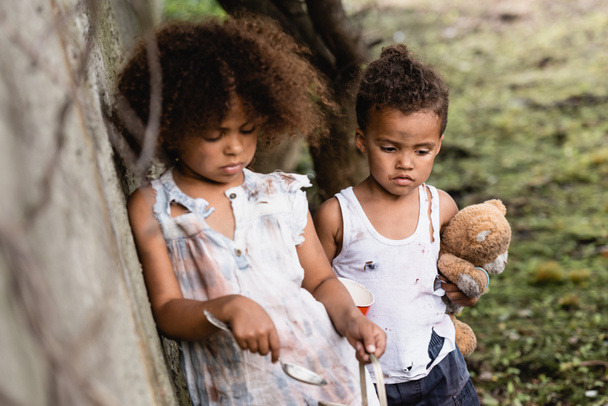 Selectieve focus van hulpeloze Afro-Amerikaanse kinderen met lepel, beker en teddybeer staan in de buurt van betonnen muur op stedelijke straat  - Foto, afbeelding