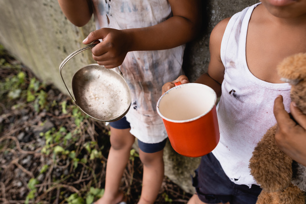 Vista ritagliata di senzatetto afro-americani bambini in possesso di tazza e piastra metallica mentre elemosina sulla strada urbana  - Foto, immagini