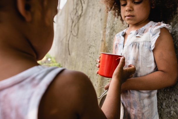 アフリカ系アメリカ人の子供の選択的な焦点は、都市の通りに汚れた服の妹に金属カップを与える  - 写真・画像