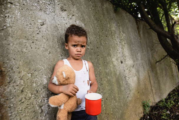 Memnuniyetsiz Afro-Amerikalı çocuk elinde kirli oyuncak ayı ve metal bardakla şehir sokağındaki beton duvarın yanında.  - Fotoğraf, Görsel