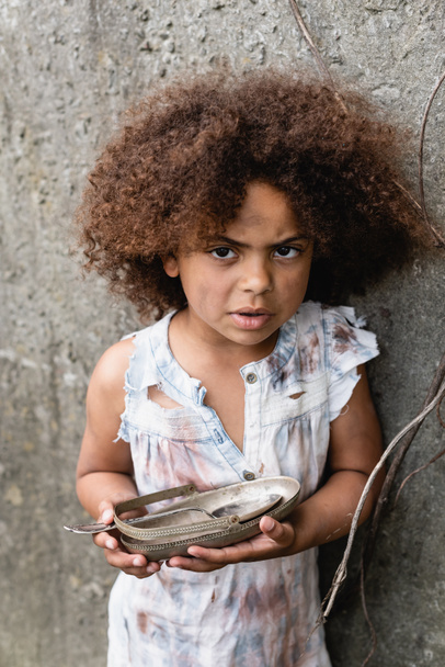 Африканский нищий американский ребенок смотрит в камеру, держа грязную тарелку и ложку возле бетонной стены в трущобах - Фото, изображение