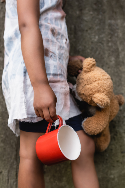 Cropped view of poor african american child holding teddy bear and metal cup on urban street  - Foto, Imagen