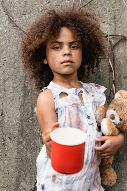Szelektív fókusz a szegény afro-amerikai gyermek kezében játékmackó és fém csésze alamizsnát a városi utcán  - Fotó, kép