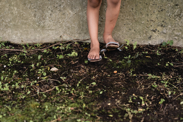 Καλλιεργημένη άποψη των ποδιών του φτωχού Αφροαμερικανού παιδιού στο έδαφος κοντά σε τσιμεντένιο τοίχο σε εξωτερικούς χώρους  - Φωτογραφία, εικόνα