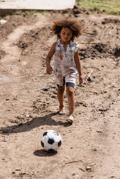 Selective focus of poor african american child playing football on dirty road on urban street  - Photo, Image