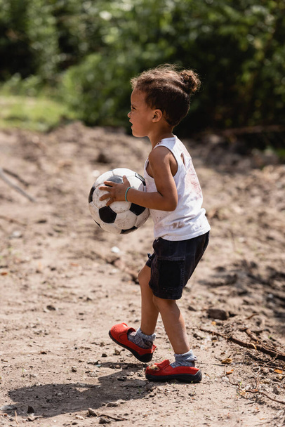 Oldalnézet a nincstelen afro-amerikai fiú kezében focilabda a piszkos úton a nyomornegyedben  - Fotó, kép
