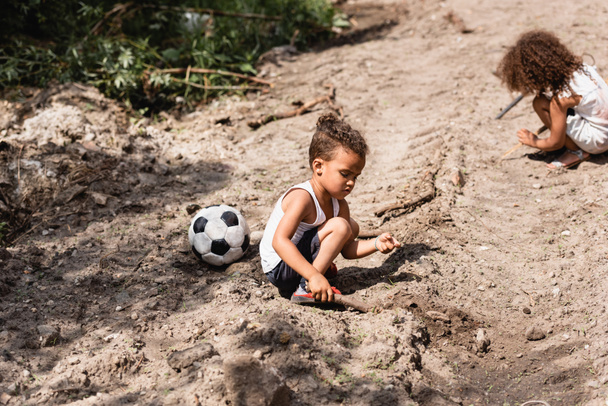 Selective focus of poor african american children playing on dirty road near football on urban street  - Photo, Image
