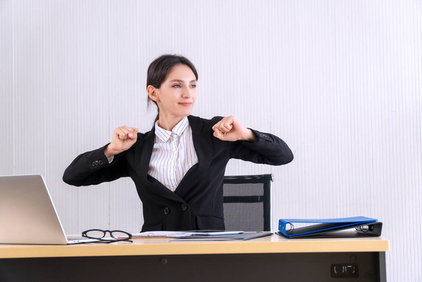 Beautiful young businesswoman stretching hands herself or exercise while sitting on table with laptop, glasses and document in break time at workplace. Office syndrome concept - Photo, Image