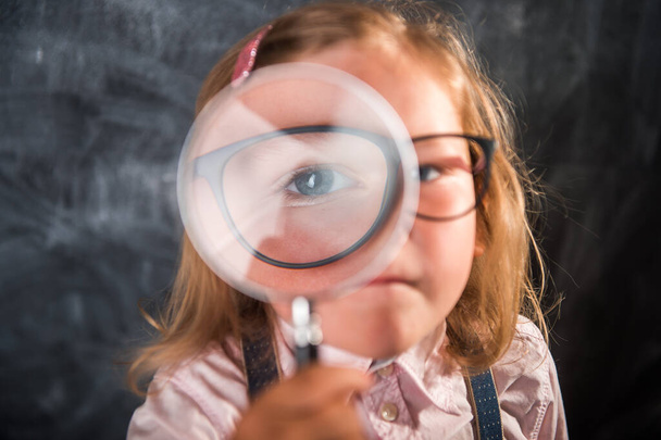 Meisje kind kijkt door een vergrootglas tegen schoolbord. Instagram filter. Terug naar school. Grappig kind gezicht. Groot oog - Foto, afbeelding