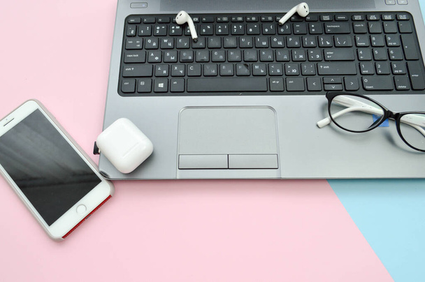 Luogo di lavoro a casa, ufficio a casa per il lavoro a distanza con un computer portatile portatile smartphone blocco note penna cuffie e occhiali su uno sfondo rosa e blu - Foto, immagini