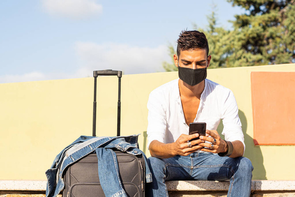 Jeune voyageur caucasien élégant avec un masque de protection COVID noir à l'aide du smartphone assis sur un banc coloré avec la veste en denim sur un sac de voyage - concept de voyage sûr Coronavirus - Photo, image