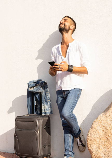 Ευτυχισμένος νεαρός κομψό καυκάσιος ταξιδιώτης άνθρωπος χαμογελώντας χρησιμοποιώντας το smartphone ακουμπά σε ένα λευκό τοίχο με ταξιδιωτική τσάντα στο έδαφος - Alwais συνδεδεμένο έννοια - Solo αρσενικό ταξιδιώτη χιλιετία - Φωτογραφία, εικόνα