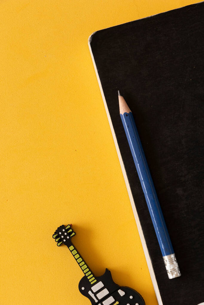 Cuaderno negro con lpiz azul al lado de un disco extraible en forma de guitarra sobre una mesa amarilla.  - Foto, Bild