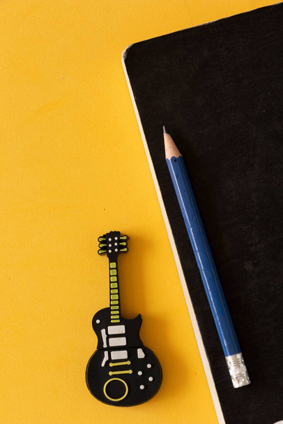 Cuaderno negro con lpiz azul al lado de un disco extraíble en forma de guitarra sobre una mesa amarilla..  - Foto, imagen