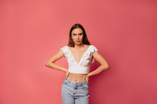 Portrait of brunette stylish lady in white top is looking at camera, arms on waist make big smile isolated on pink bright background - Фото, изображение