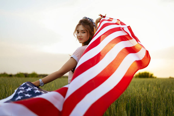 young happy beautiful caucasian teenager girl in pink top and jeans shorts with national american flag on her in green wheat field at sunset view from the back. Independence day - Photo, Image