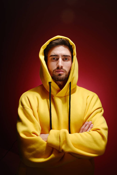 A young man of 25-30 years old in a yellow sweatshirt is standing against a red wall.  - Photo, Image
