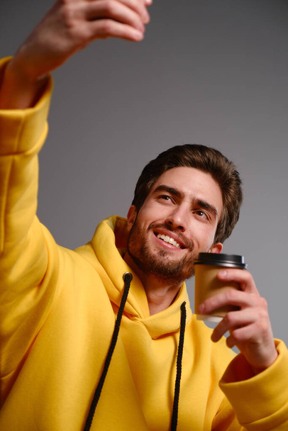 Young smiling guy in a yellow sweatshirt with a disposable paper glass of coffee in hand makes selfie on a smartphone - Photo, Image