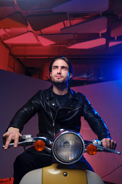 A young man of 25-30 years in in a leather jacket and black jeans posing while sitting on a moped on a purple background. - Foto, imagen